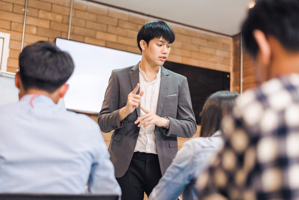 business-cooperation-young-asian-male-coach-or-speaker-make-flip-chart-presentation-to-diverse-businesspeople-at-meeting-in-office-male-tutor-or-trainer-present-project-to-diverse-colleagues